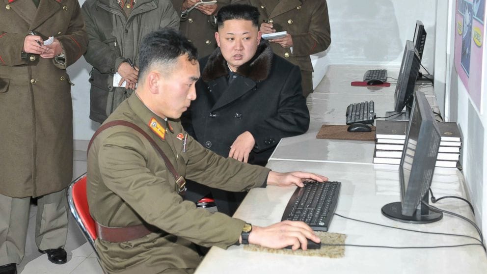 a photo of a North Korean soldier, in full uniform, sat at a computer looking nervous as hell, as Kim Jong Un sits next to him in a black coat, looking at the computer screen.