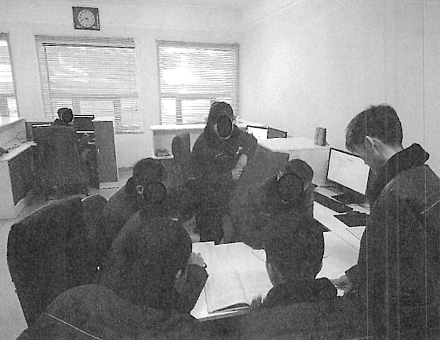 a team of North Koreans looking over books and textbooks working at a table, surrounded by computers, in a lab/apartment where the North Korean IT workers' scheme is being run from; the photo is monochrome and grainy.