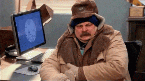 an animated GIF of Nick Offerman, playing Ron Swanson, sat at his computer desk layered up with jackets, gloves, and several hats, seemingly freezing cold.