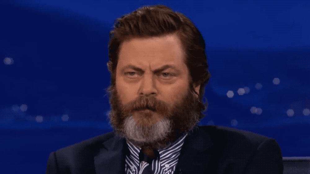 A close up of a beareded Nick Offerman looking very serious and angry, then suddenly bursting into a happy smile and a giggle.