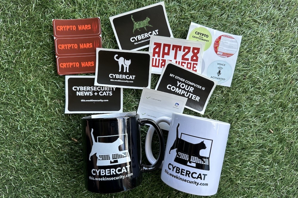 A collection of This Week in Security themed stickers and two mugs, one in black, one in white, on a grassy background.