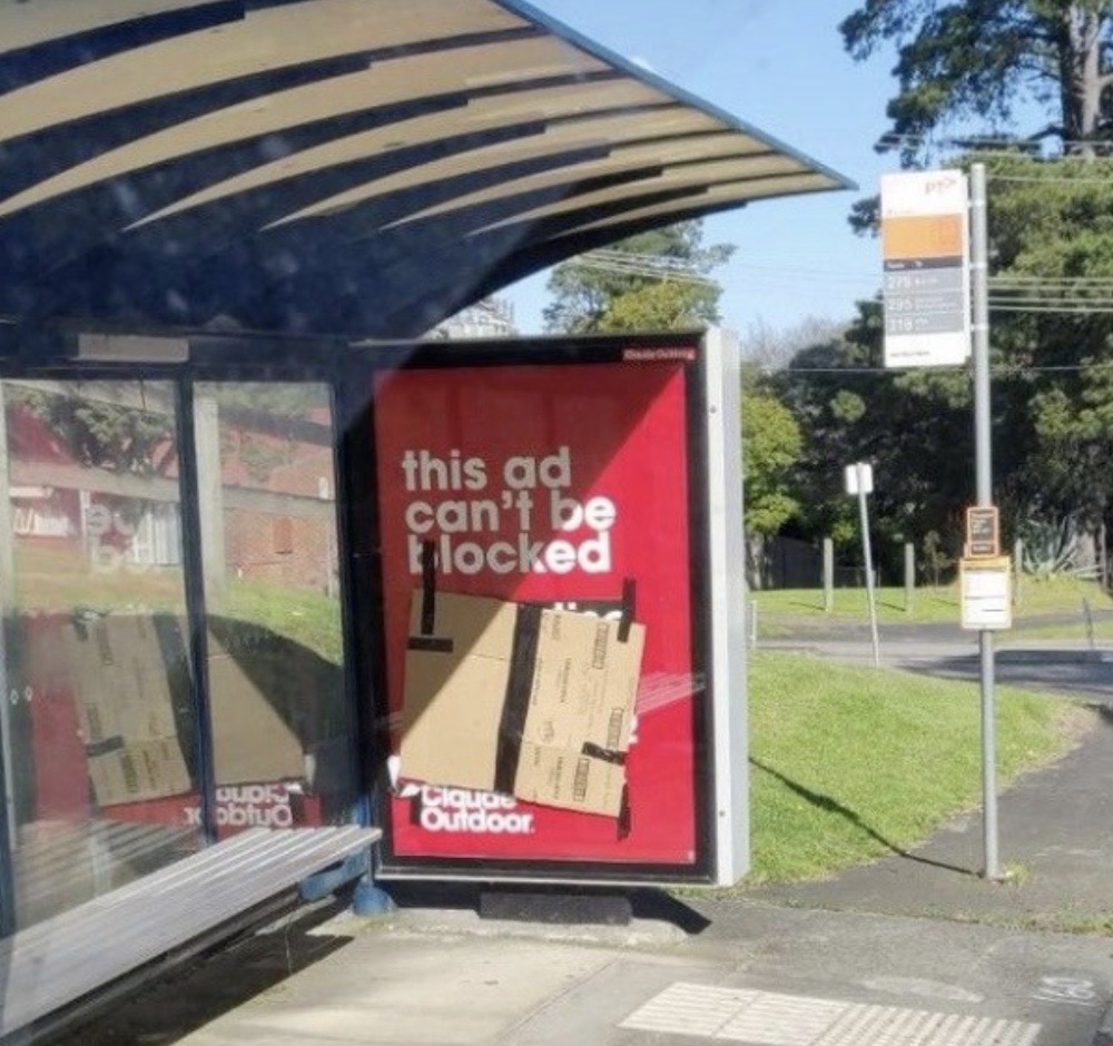 A photo of a real-world bus stop with an advertisement that says, "this ad can't be blocked," but someone has taped a large cardboard box to the ad, obscuring its text.