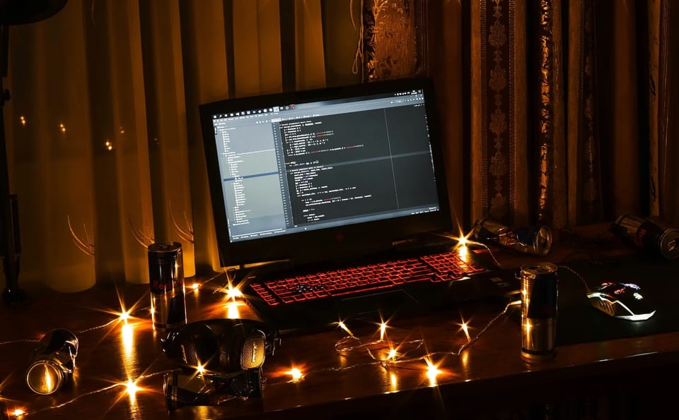 Photo of a programmer's laptop with a red-backlit keyboard, with many energy drinks and holiday twinkly lights, in a darkened room.